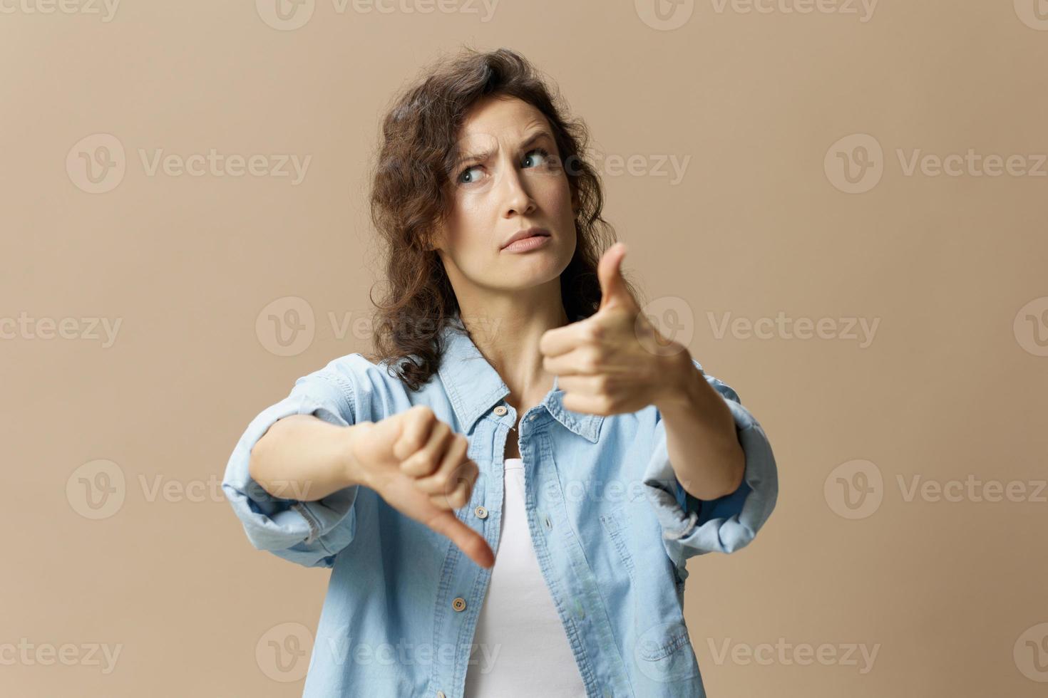 Confused curly beautiful female in jeans casual shirt can not make decision show thumbs up down looks up posing isolated on over beige pastel background. People Lifestyle emotions concept. Copy space photo