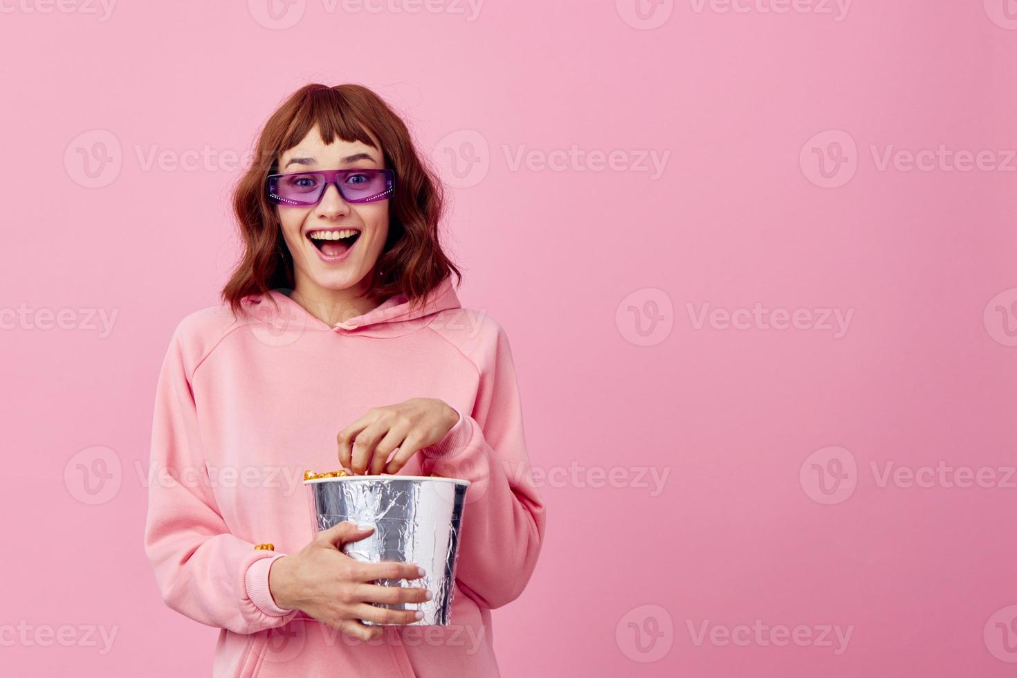 disfruté contento sonriente linda pelirrojo dama en rosado capucha Gafas de sol con palomitas de maiz posando aislado en terminado rosado estudio antecedentes. Copiar espacio bandera. Moda cine película concepto. entretenimiento oferta foto