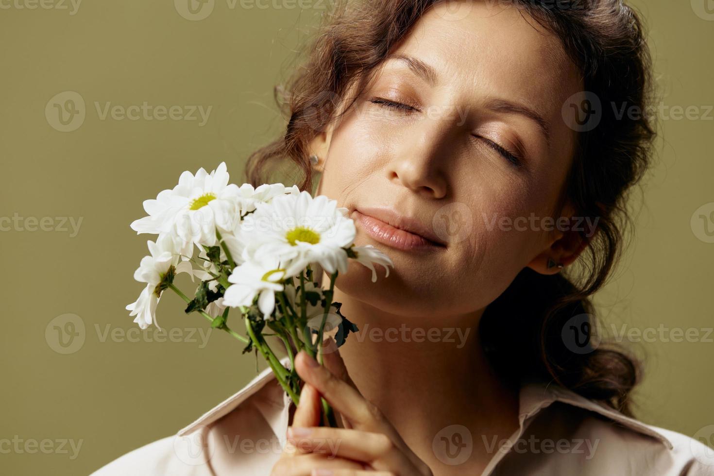 Close up portrait of cheerful curly female in linen casual shirt hold chamomiles enjoy flowers smell close eyes posing isolated on olive green pastel background. Nature is beauty concept. Copy space photo