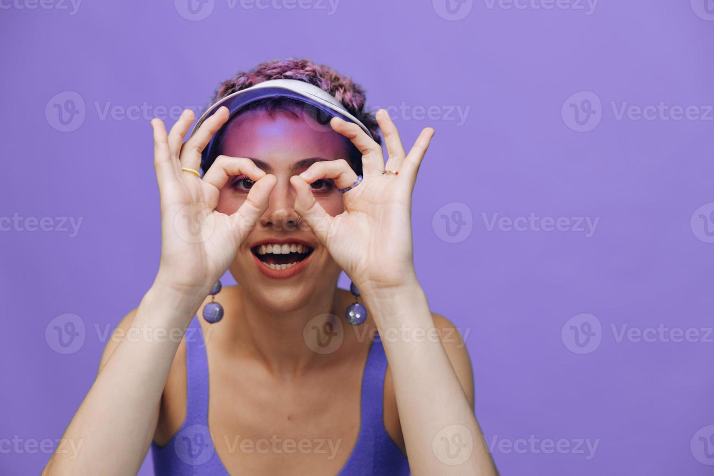Portrait of a sporty fashion woman posing smiling with teeth at the camera with stylish earrings in her ears in a purple yoga tracksuit and a transparent cap on a purple monochrome background photo