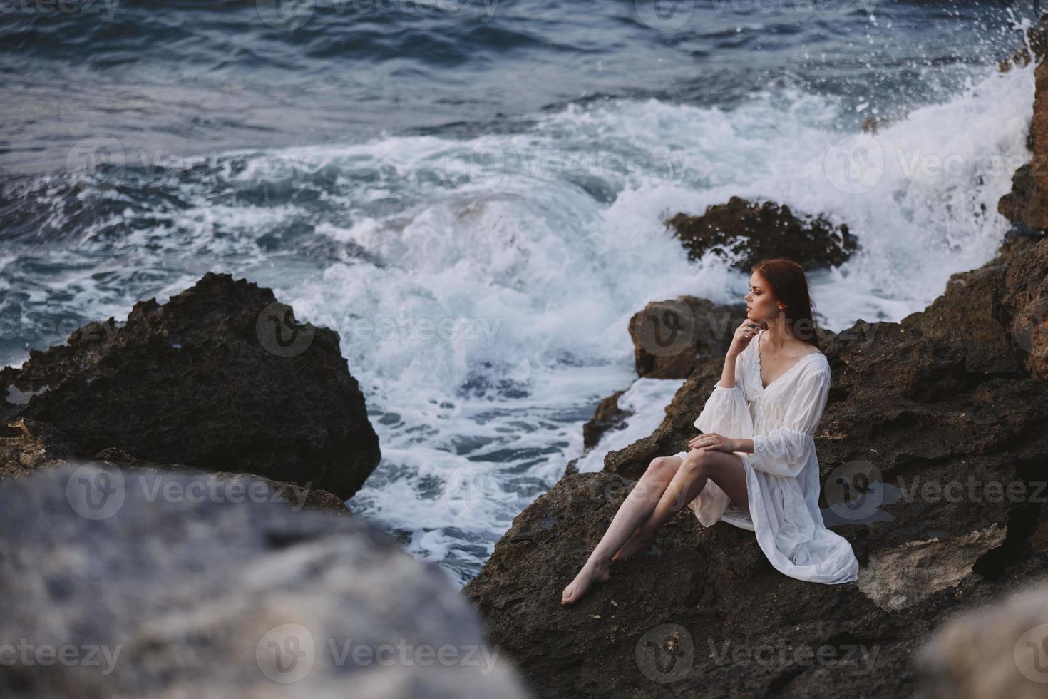 hermosa mujer en un blanco Boda vestir se sienta en el piedras por el Oceano pensativo Mira foto