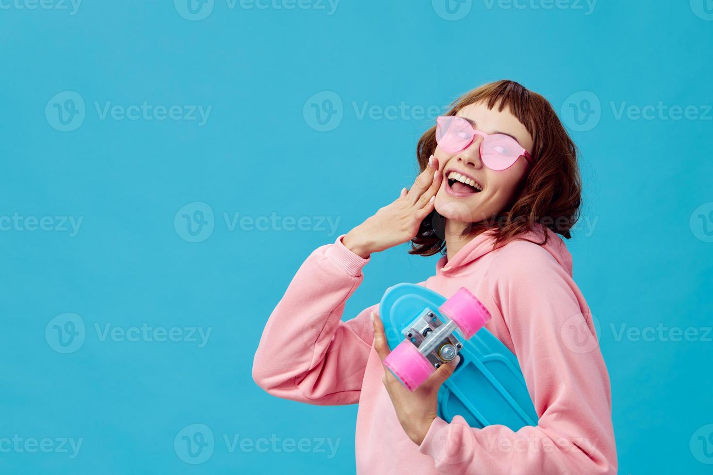 Happy cheerful pretty redhead lady in pink hoodie sunglasses with penny board posing isolated on blue studio background. Copy space Banner Offer. Fashion Cinema. Holiday activity concept photo