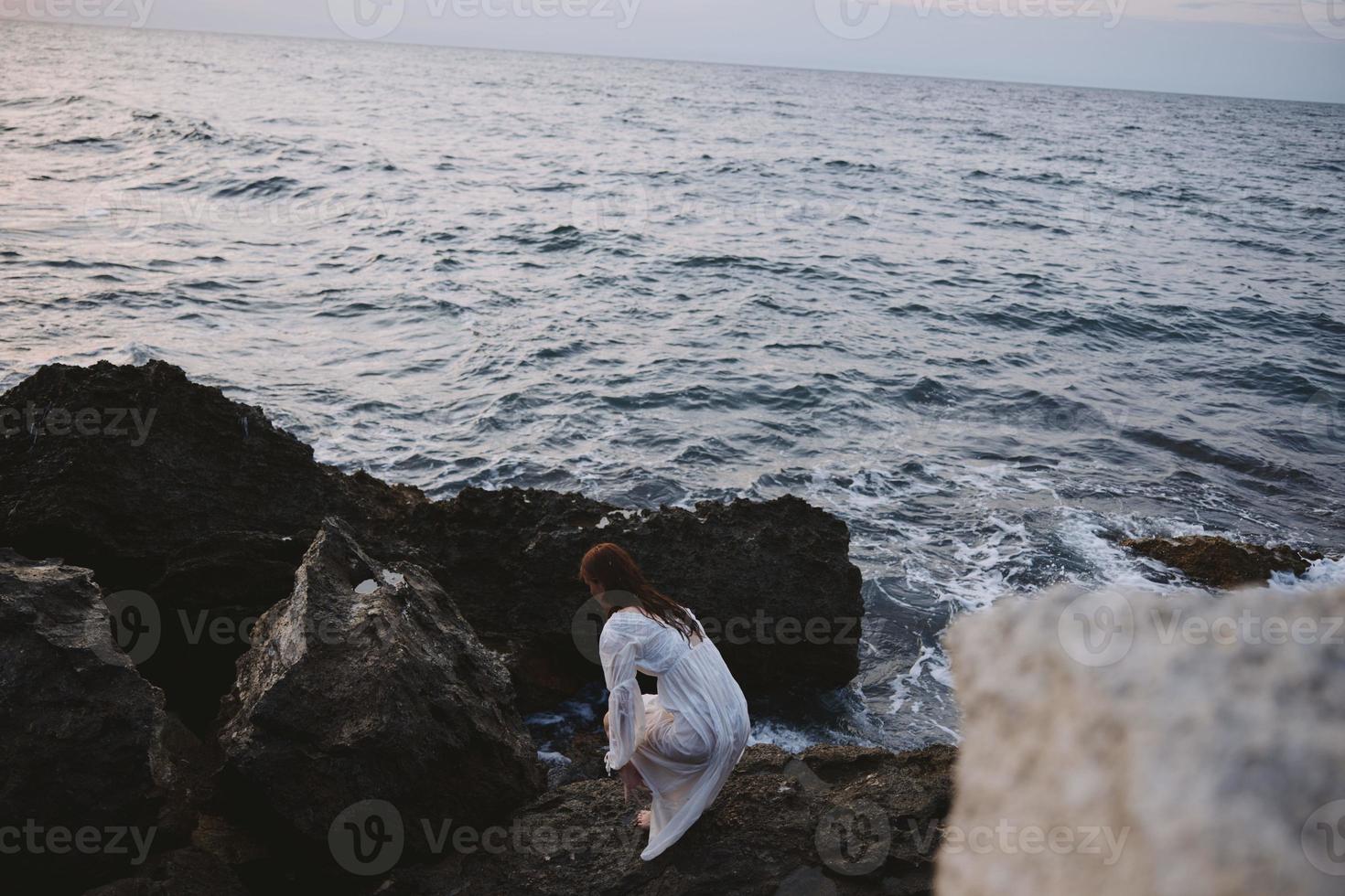 barefoot woman in a white dress with wet hair stands on a cliff photo