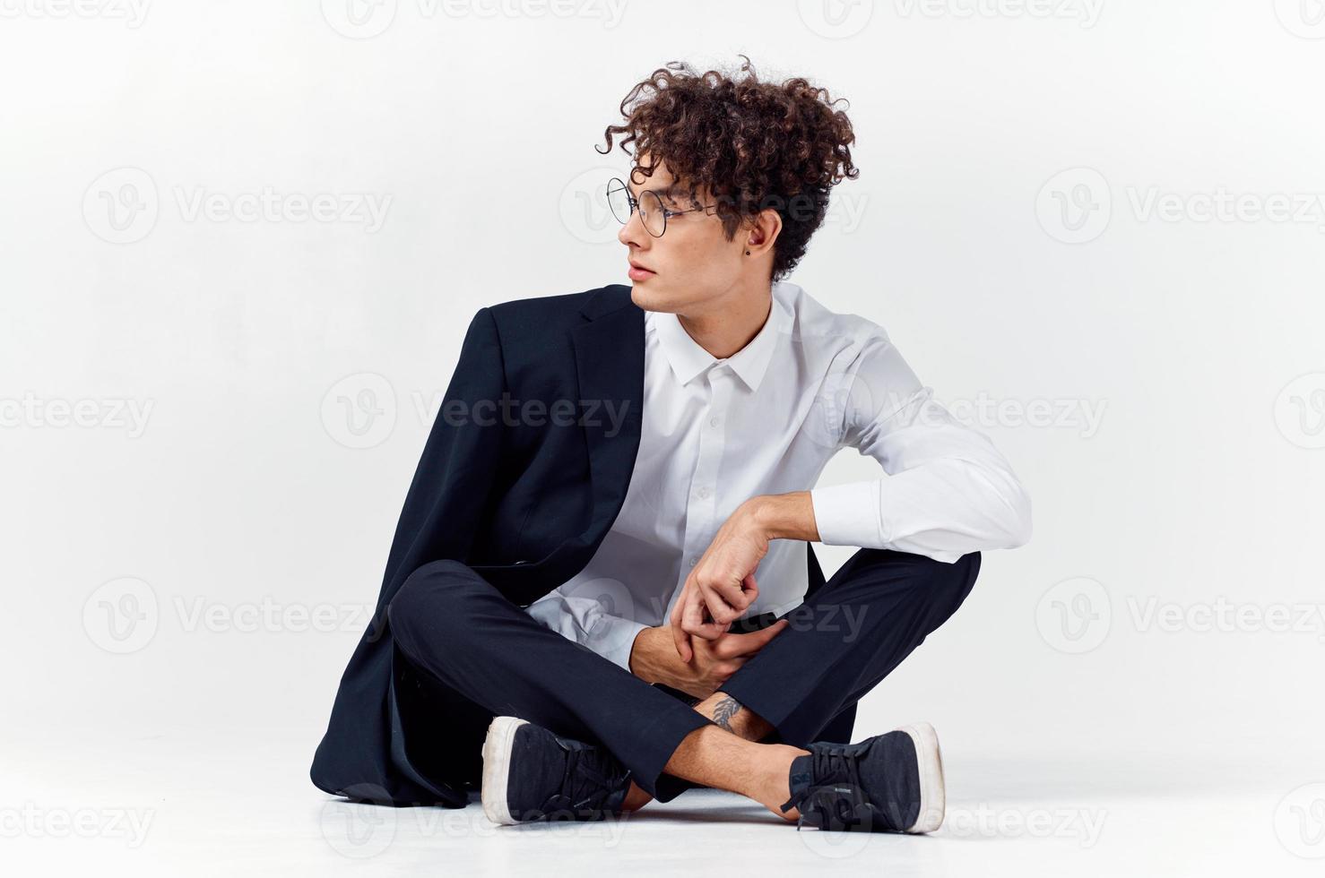 curly-haired guy in fashionable clothes and sneakers sits on the floor in a bright room in full growth photo