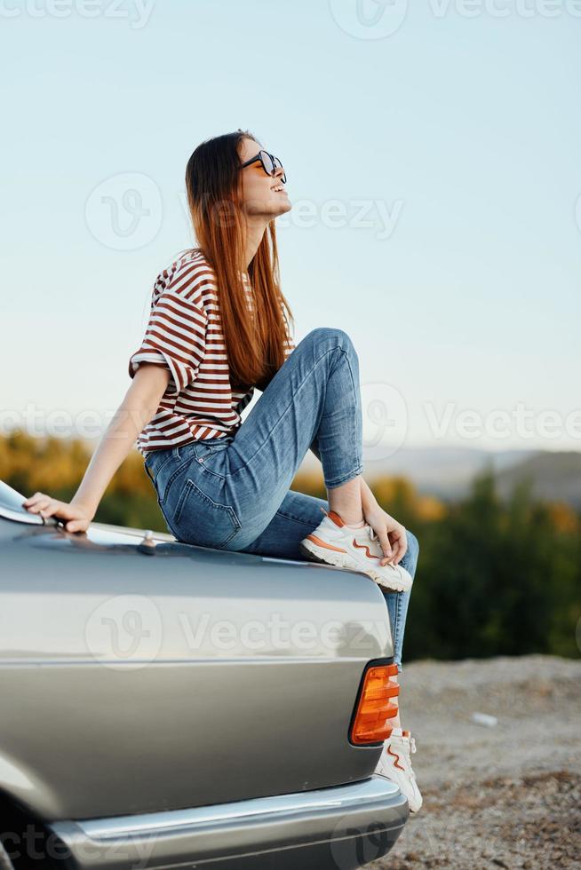 un Moda mujer en elegante anteojos, un a rayas camiseta y pantalones se sienta en el maletero de un coche y mira a el hermosa naturaleza de otoño foto