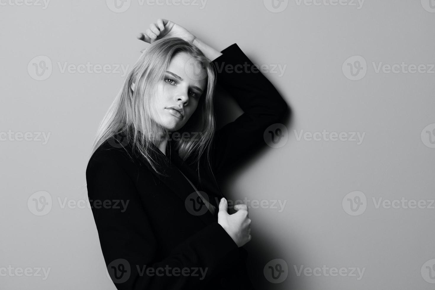 Serious young blonde woman leans on the studio wall holding hand overhead looks at camera posing isolated in black jacket photo