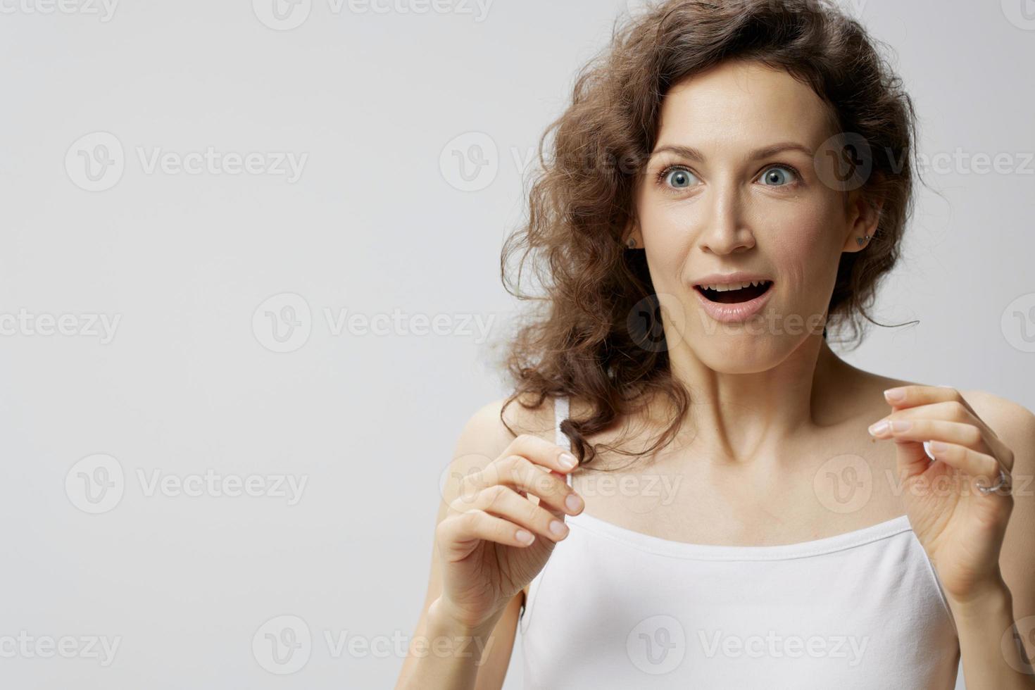 Confused shocked curly beautiful woman in basic white t-shirt raise hands up open mouth posing isolated on over white background. People Emotions Lifestyle. I can not believe it concept. Copy space photo