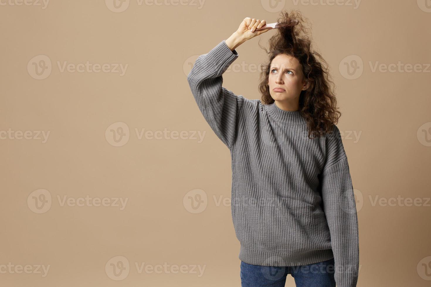 Funny thoughtful curly beautiful female in gray casual sweater with hairbrush comb pulls curls up posing isolated on over beige pastel background. Problematic unruly damaged hair concept. Copy spaceFunny thoughtful curly beautiful female in gray casual sw photo