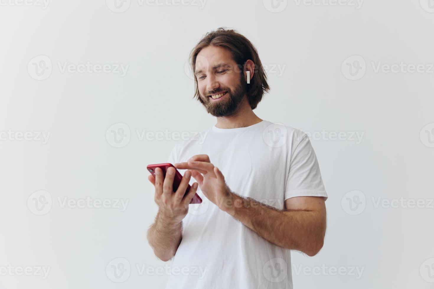 A man with a beard and long hair in a white T-shirt and blue jeans looks at his phone flipping through an online social media feed with headphones in his ears against a white background photo