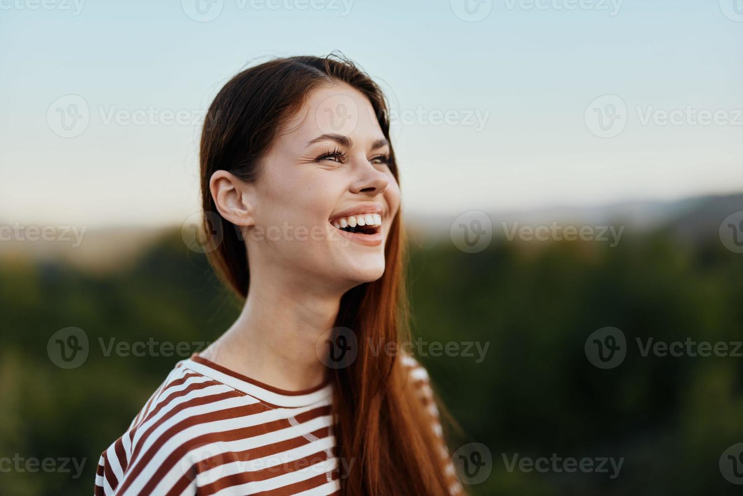 Close-up portrait of a young woman with a beautiful smile with teeth in a striped t-shirt against the background of trees photo