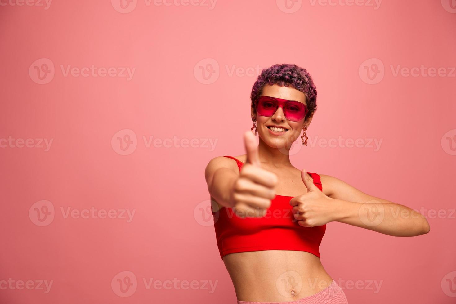 Fashion portrait of a woman with a short haircut in colored sunglasses with unusual accessories with earrings smiling on a pink bright background showing thumbs up at the camera photo