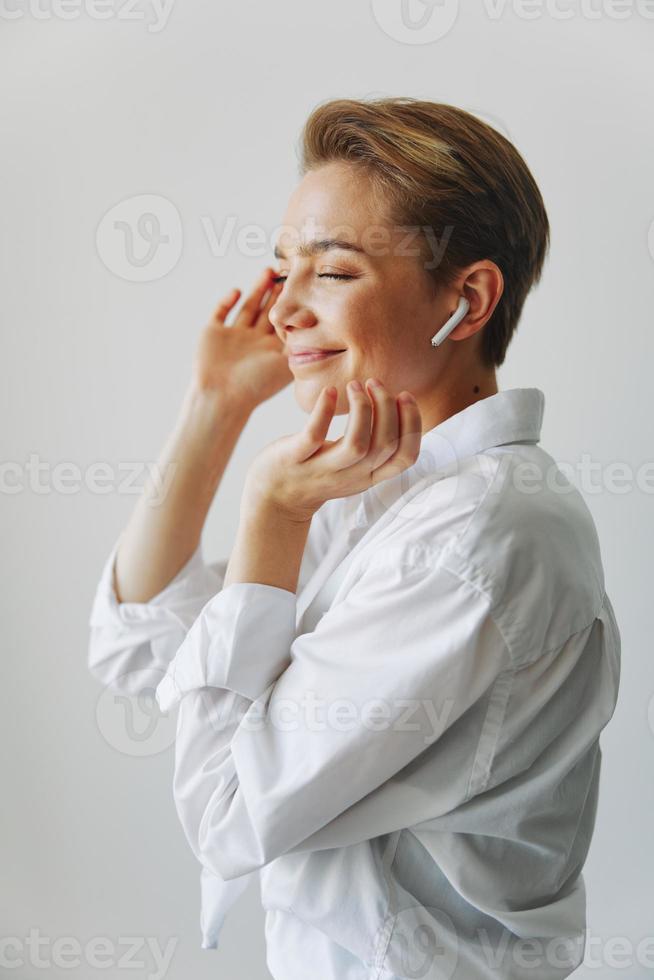 Young woman teenager listening to music with infertile headphones and dancing home, grinning with teeth with a short haircut in a white shirt on a white background. Girl natural poses with no filters photo