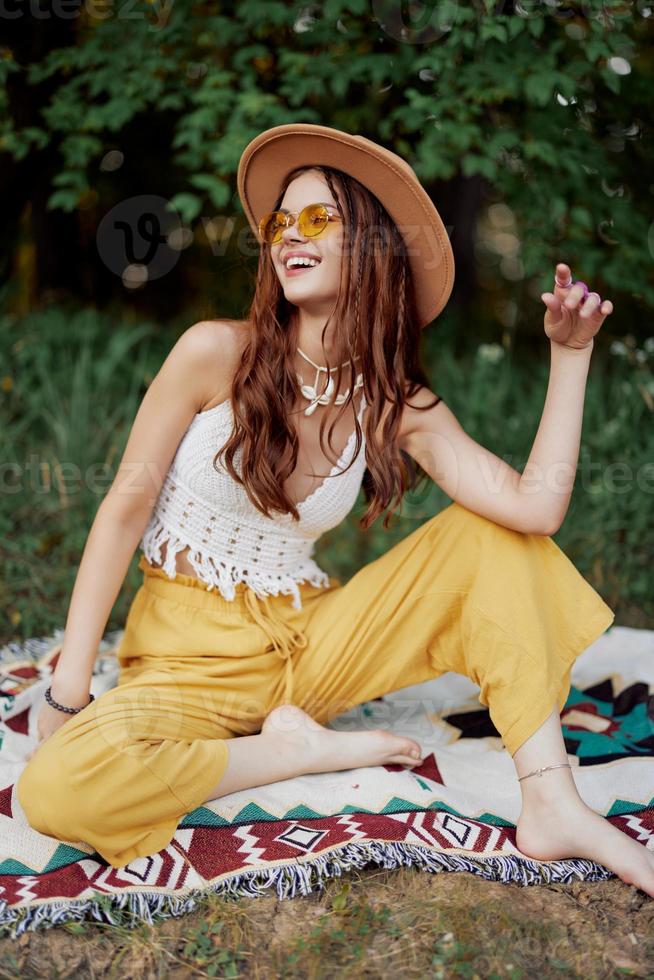 Hippie woman smiling in eco clothing yellow pants, white knit top, hat and yellow glasses sitting on plaid in park watching sunset, lifestyle camping trip photo