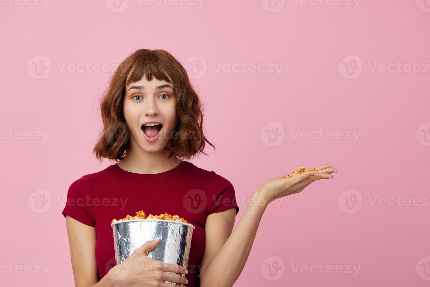 emocionado conmocionado linda pelirrojo dama en rojo camiseta con palomitas de maiz Listo para película noche posando aislado en terminado rosado estudio antecedentes. Copiar espacio bandera. Moda cine concepto. entretenimiento oferta foto