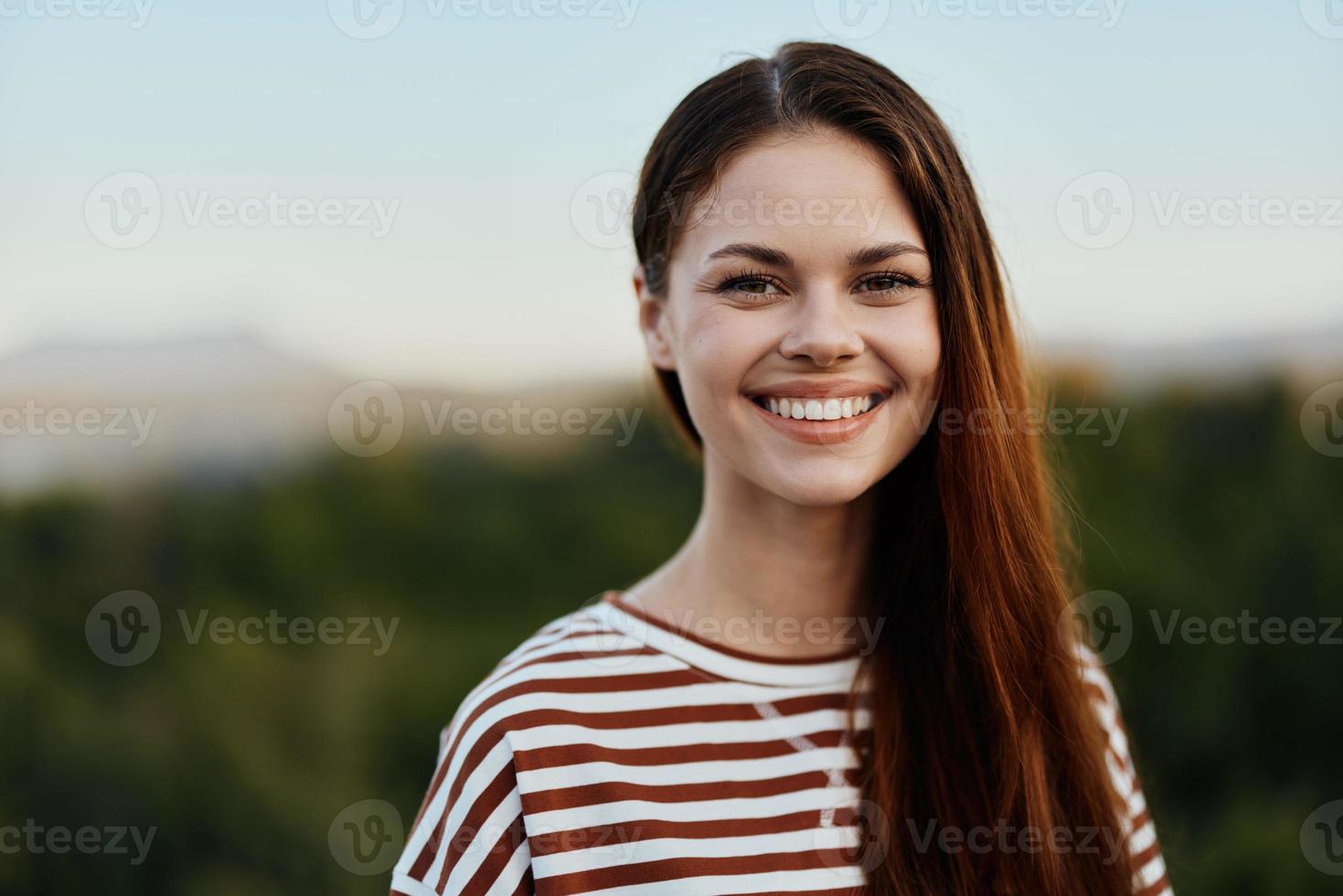 Close-up portrait of a young woman with a beautiful smile with teeth in a striped t-shirt against the background of trees photo