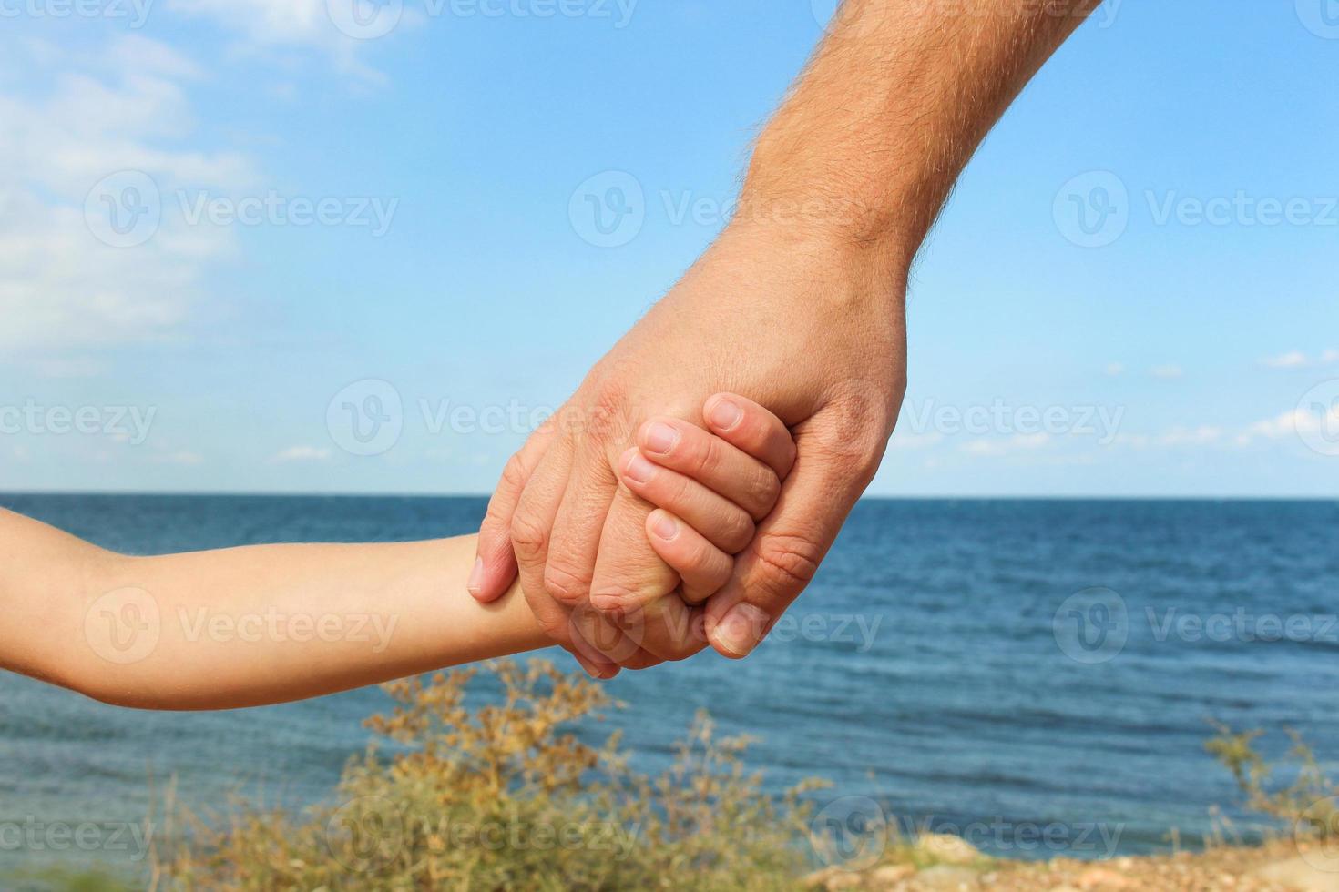 Man holds hand of child on background of sea and sky Concept of love, care, friendship, trust in family. photo