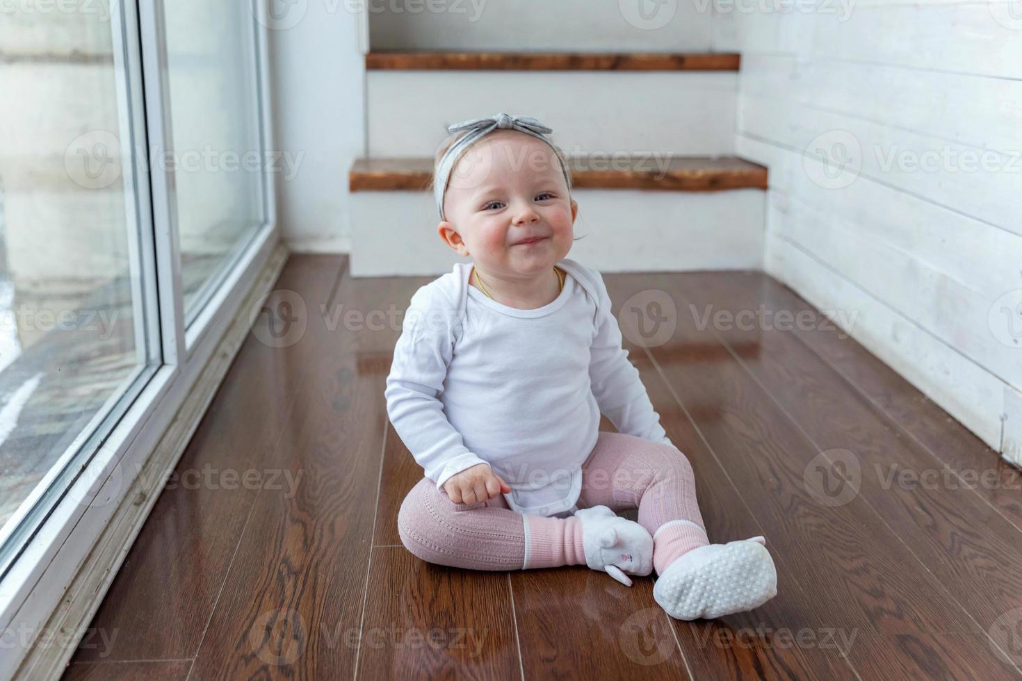 Little crawling baby girl one year old siting on floor in bright light living room near window smiling and laughing photo