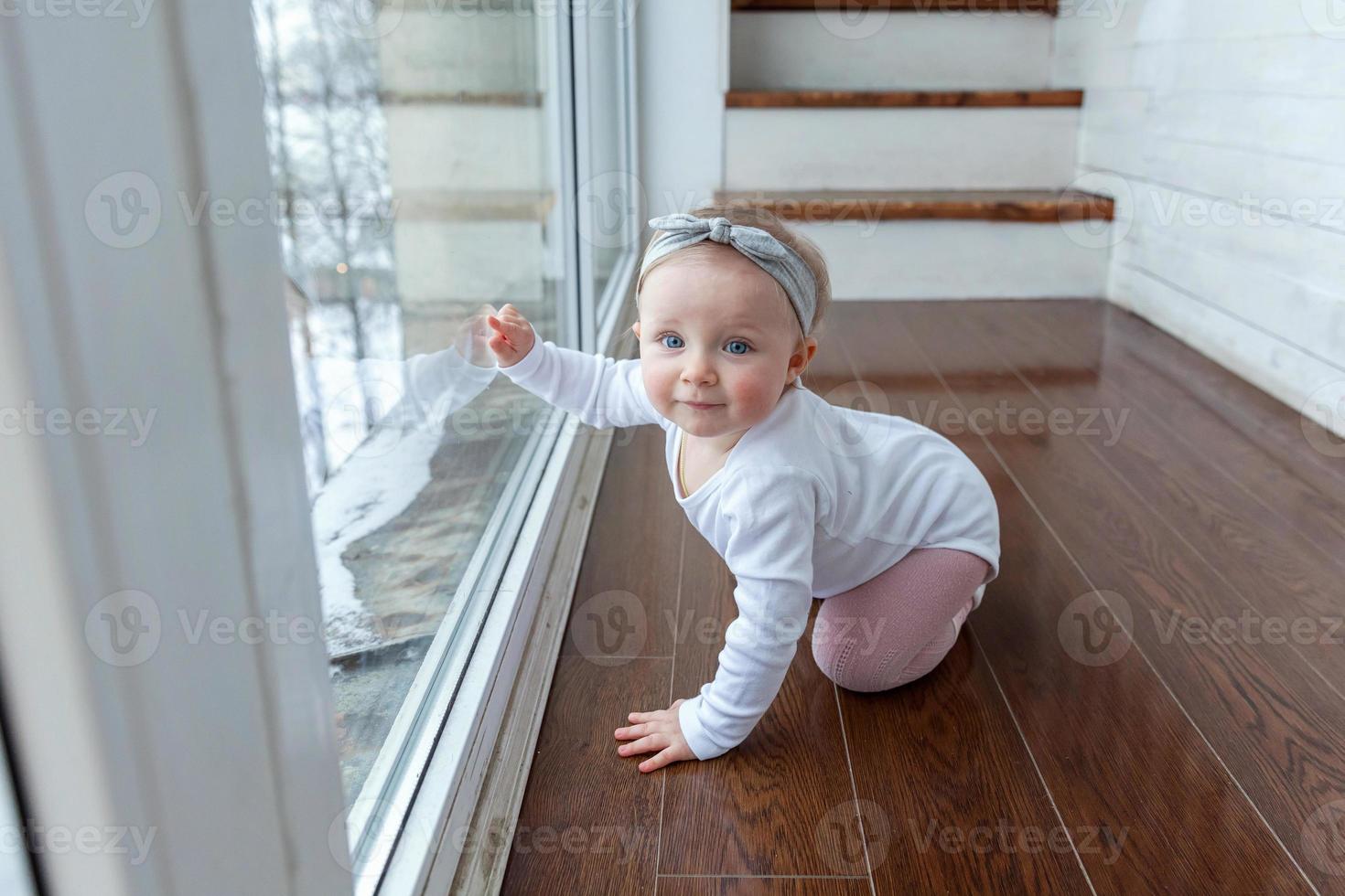 Little crawling baby girl one year old siting on floor in bright light living room near window smiling and laughing photo