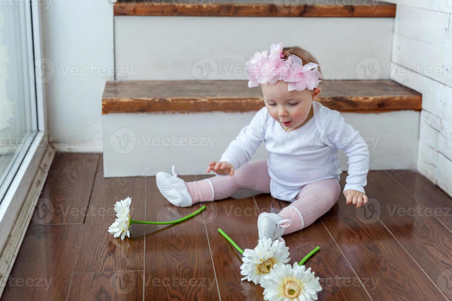 Little baby girl wearing spring wreath siting on floor in bright light living room near window and playing with gerbera flowers photo