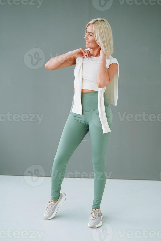 Young smiling blonde woman in sportswear doing stretching on background of grey wall photo