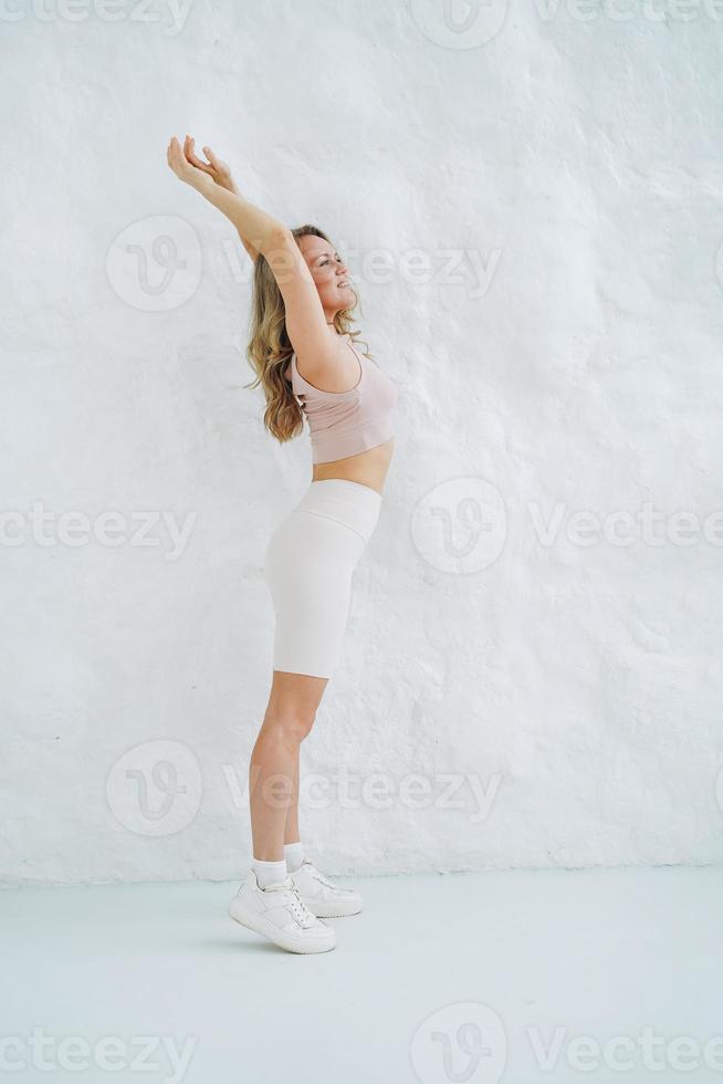 Young smiling blonde woman in sportswear doing stretching on background of white wall photo