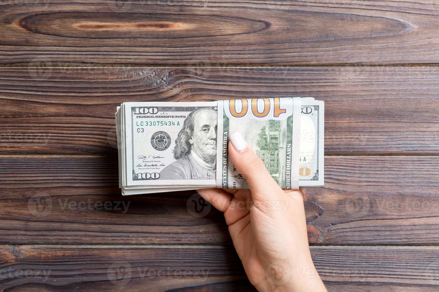 Top view of female hand holding a pack of one hundred dollar bills on wooden background. Wage and salary concept photo