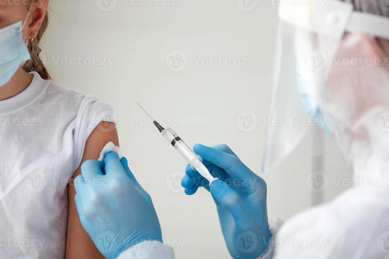 un médico en un protector traje hace el niño en un médico máscara un hombro Disparo . coronavirus vacunación. covid-19 vacuna. médico vacunar niño. niños a clínica. foto