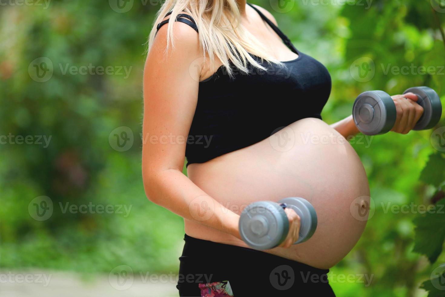 Close-up belly of a pregnant girl. A pregnant woman goes in for sports on the street, holding a young dumbbell in her hands. photo