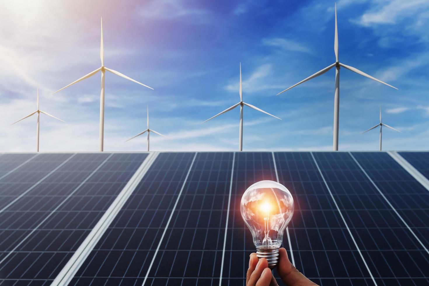hand holding lightbulb with solar panel and wind turbine background. clean energy in nature concept photo