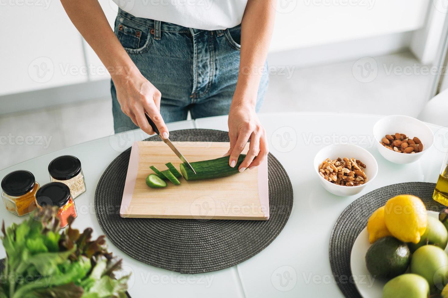 Young slim woman in white t-shirt and blue jeans cooking healthy food with cucumber in kitchen at home photo