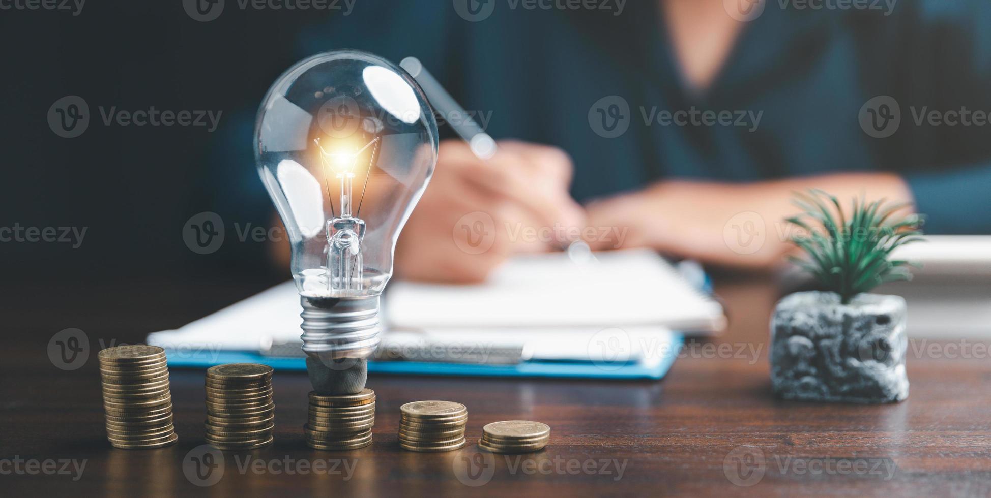 Businesswoman with lightbulb on coins and using calculator to calculate and money stack. Save energy and money with accounting finance in office concept. Idea of energy saving planning in home. photo