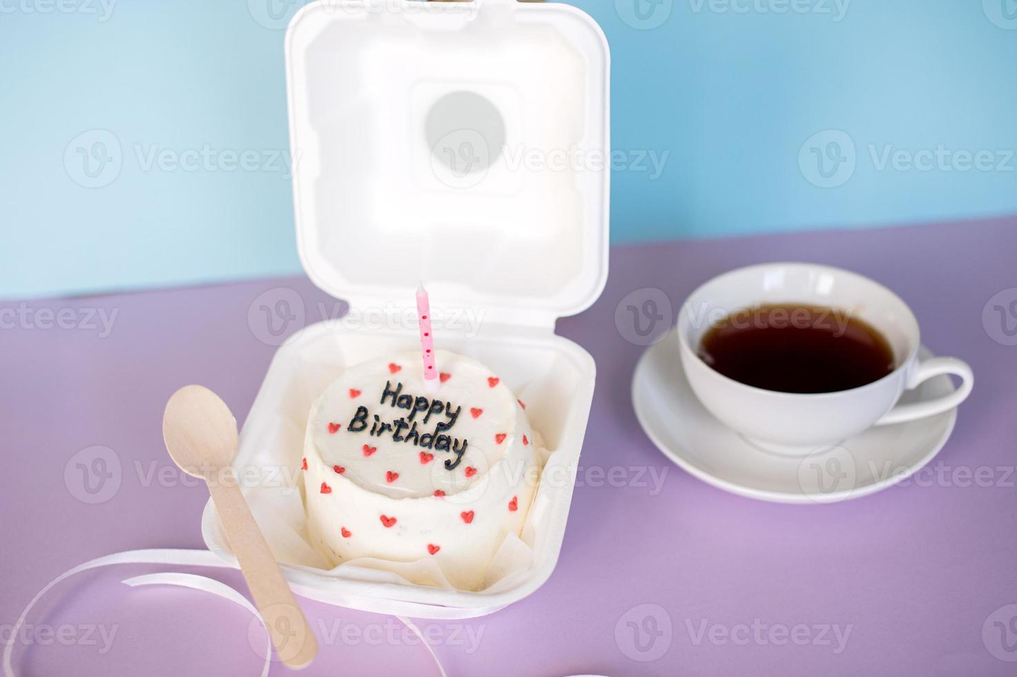 A small bento cake in eco-packaging with a wooden spoon and a candle next to a cup of tea on a purple and blue background photo