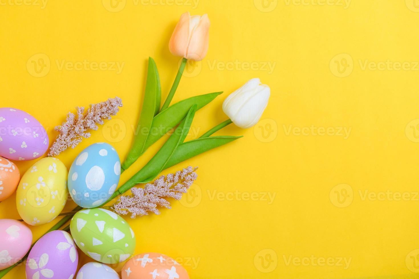 Happy Easter holiday greeting card design concept. Colorful Easter Eggs and spring flowers on yellow background. Flat lay, top view, copy space. photo