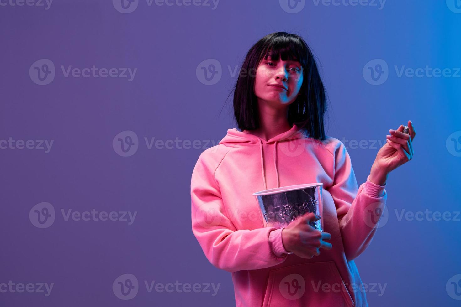 Happy awesome brunet woman in pink hoodie with popcorn look at camera posing isolated in blue violet color light studio background. Neon party Cyberpunk Cinema concept. Copy space photo