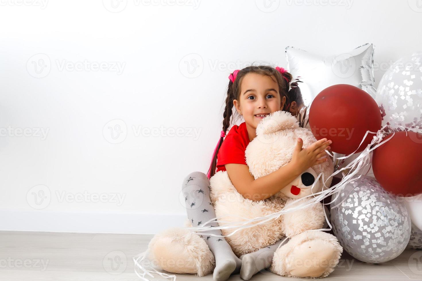 Cute kid little girl posing with red heart shaped balloons and a teddy bear isolated on white. Children fashion photo