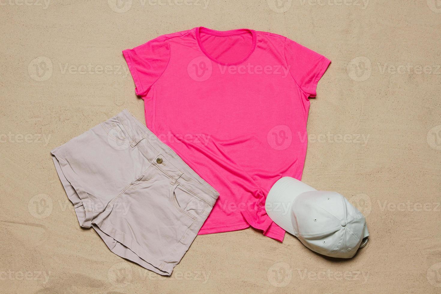 Sand beach texture background. Mockup pink summer t-shirt outfit copy space. Blank template woman shirt Top view. Summertime accessories hat, jeans. Flat-lay closeup tshirt on seashore. Beachtime photo