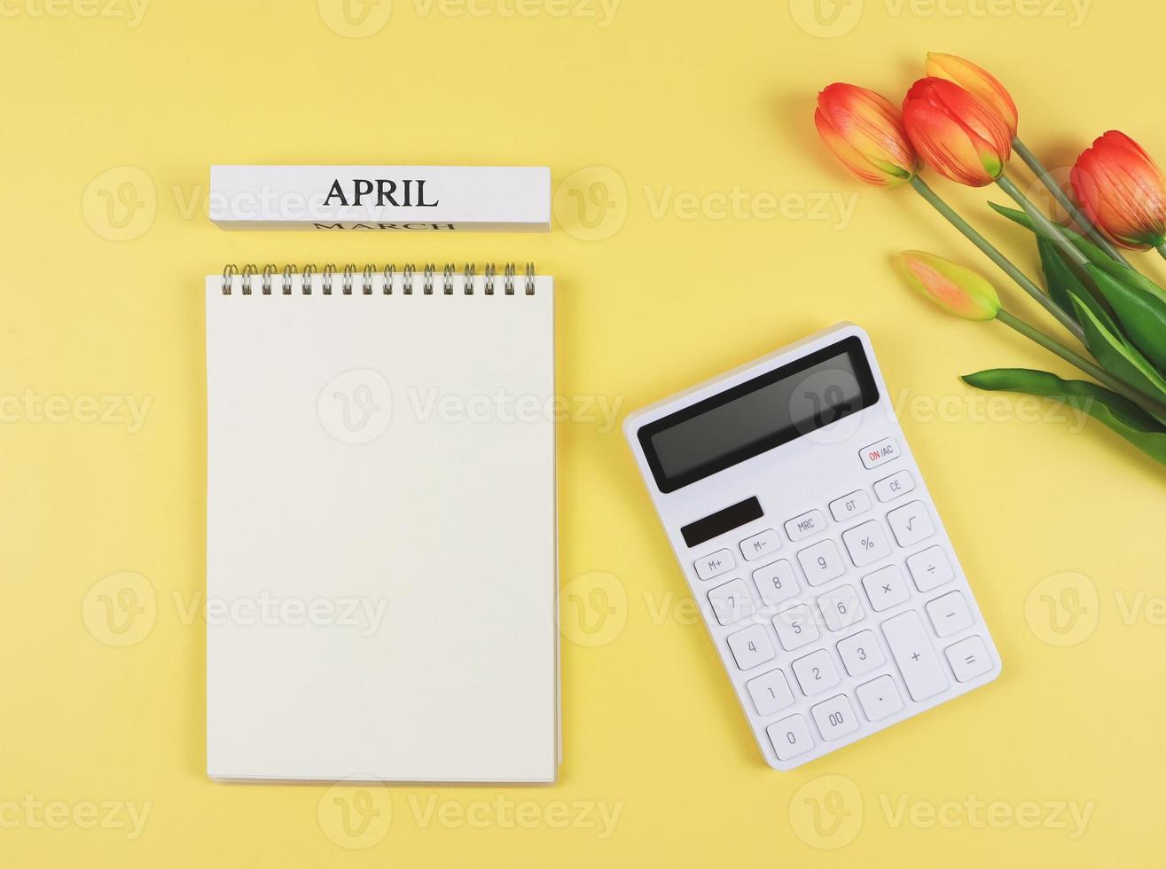 plano diseño de abrió blanco página cuaderno con de madera calendario abril, blanco calculadora y tulipanes en amarillo antecedentes. mensual financiero plan concepto. foto