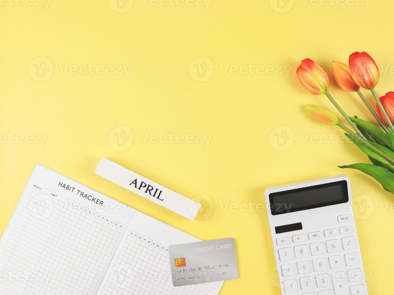 flat layout of Habit tracker book,  white calculator, credit card, wooden calendar April and tulips on yellow background. photo