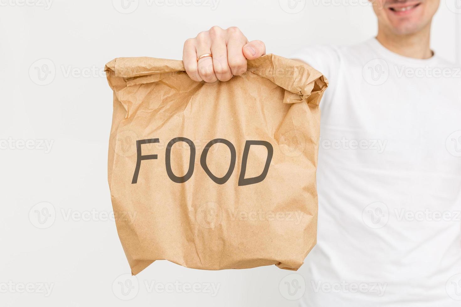 Young man holding paper bag, close-up. Wearing t-shirt, light grey background. Food delivery. photo