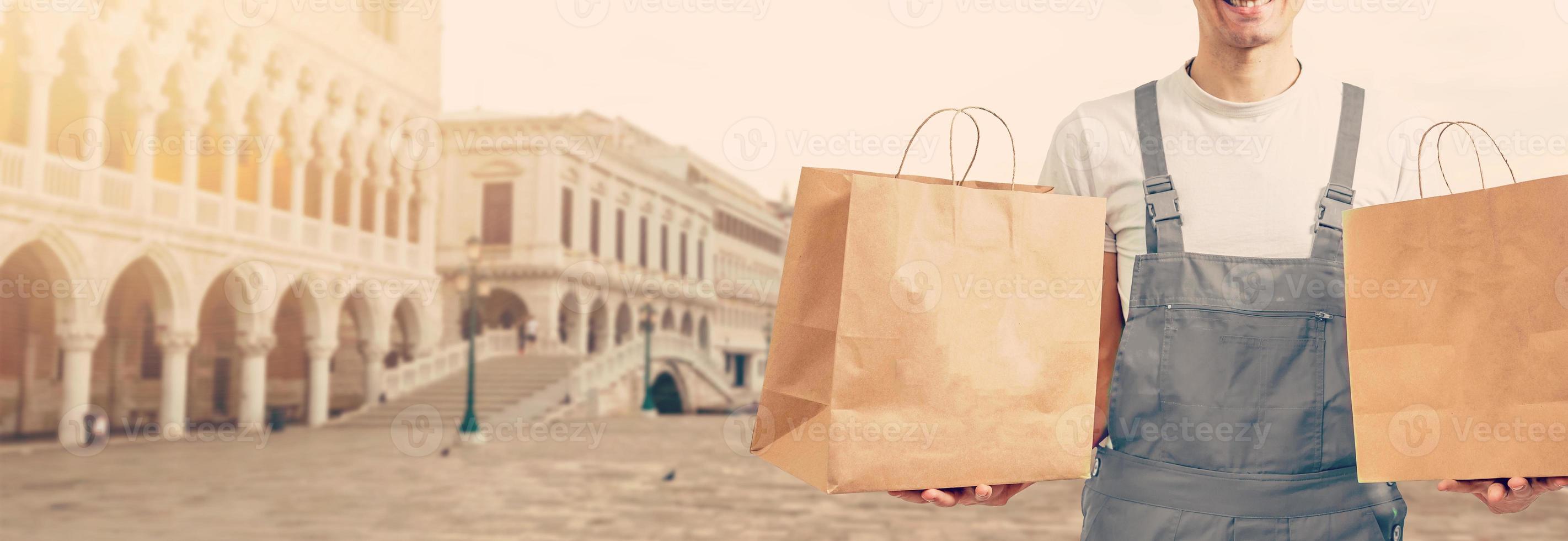 Young man holding paper bag, close-up. Wearing t-shirt, light grey background. Food delivery. photo