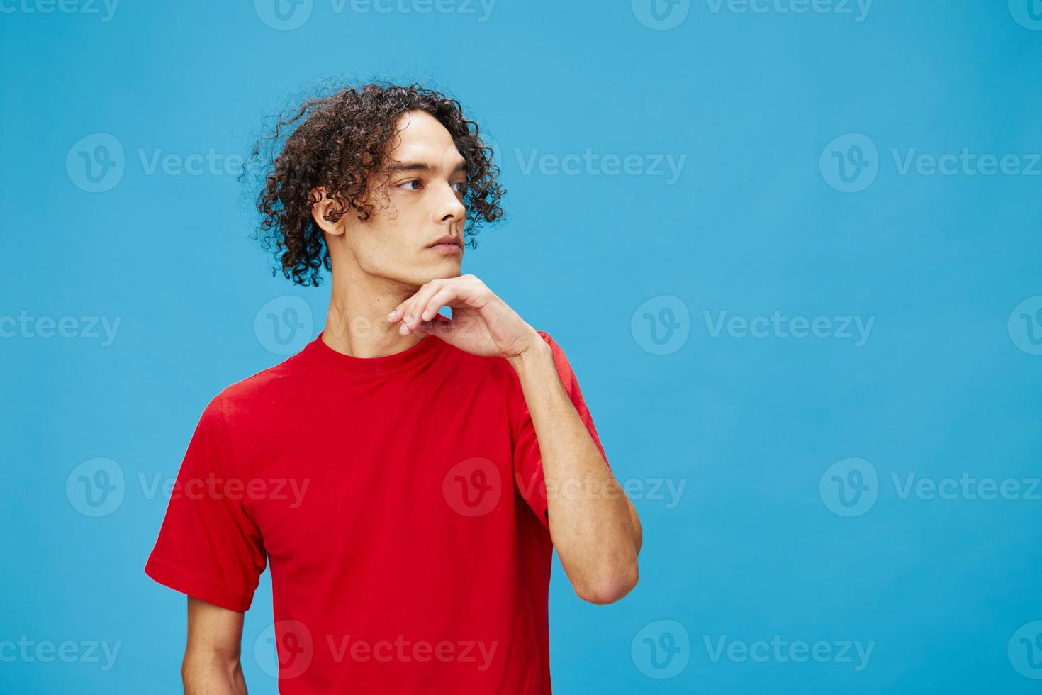 Pensive thoughtful unhappy young curly tanned Caucasian guy in basic red t-shirt posing isolated on over blue background. Lifestyle and Emotions concept. Good offer with copy space for ad photo