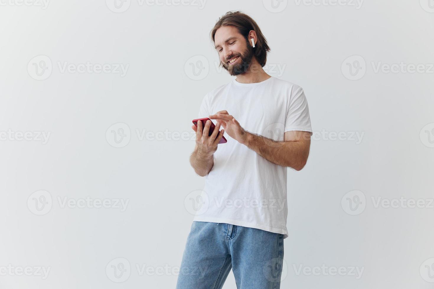 A man with a beard and long hair in a white T-shirt and blue jeans looks at his phone flipping through an online social media feed with headphones in his ears against a white background photo