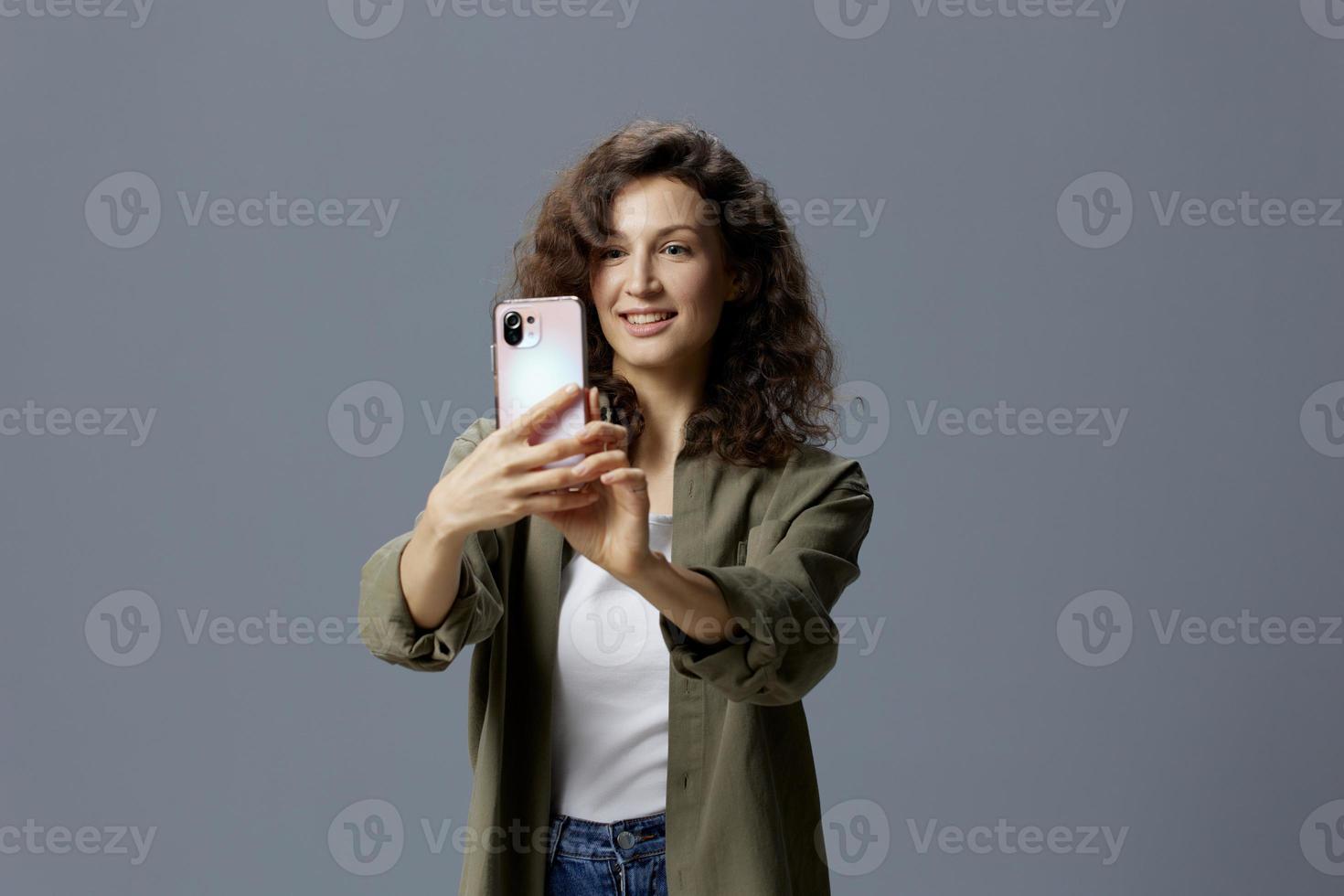 Happy smiling curly beautiful woman in casual khaki green shirt doing selfie video call using phone posing isolated on over gray blue background. Social Media Influencer concept. Copy space photo