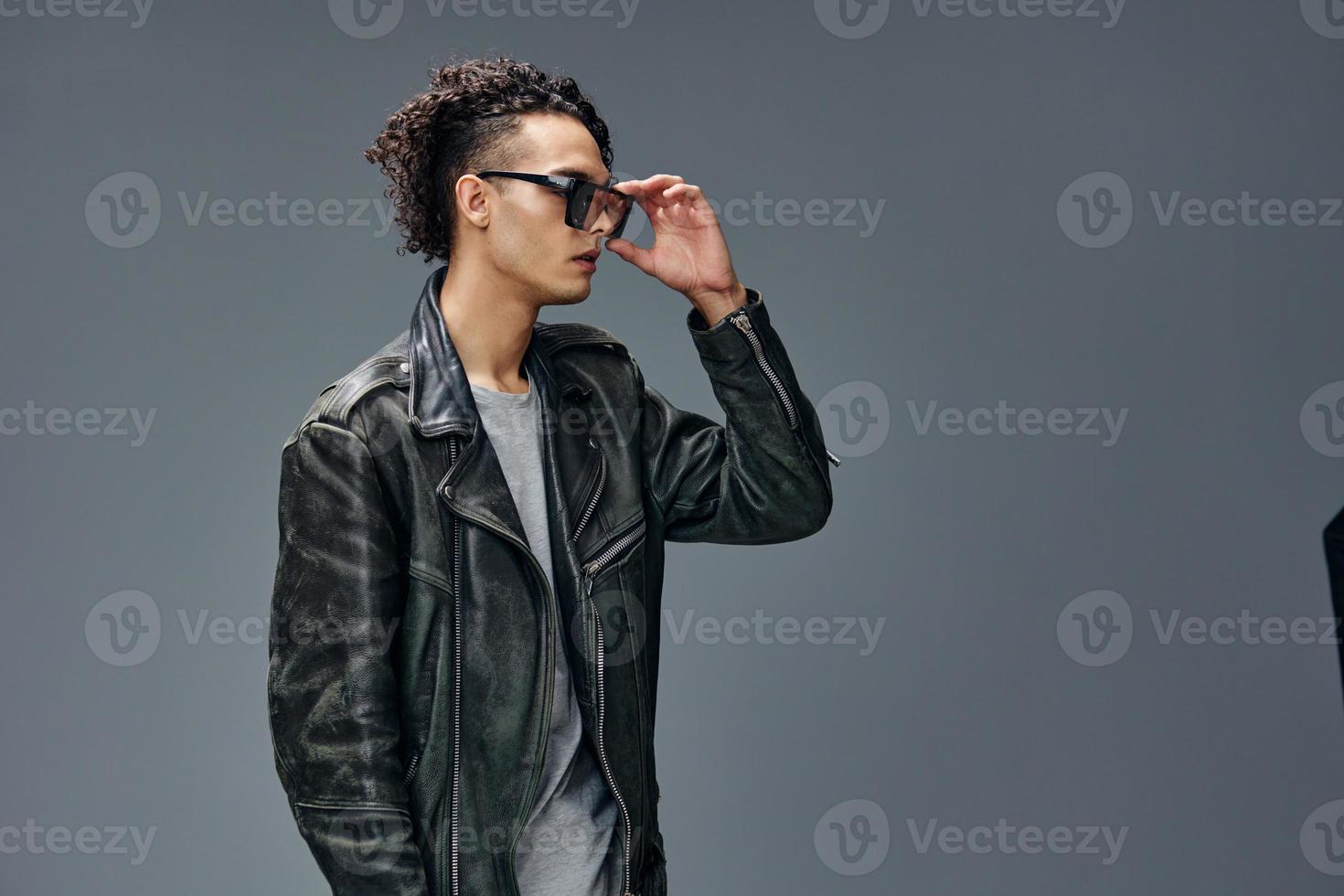 Stylish tanned curly man leather jacket mirror trendy eyewear looks aside posing isolated on gray studio background. Cool fashion offer. Huge Seasonal Sale New Collection concept. Copy space for ad photo