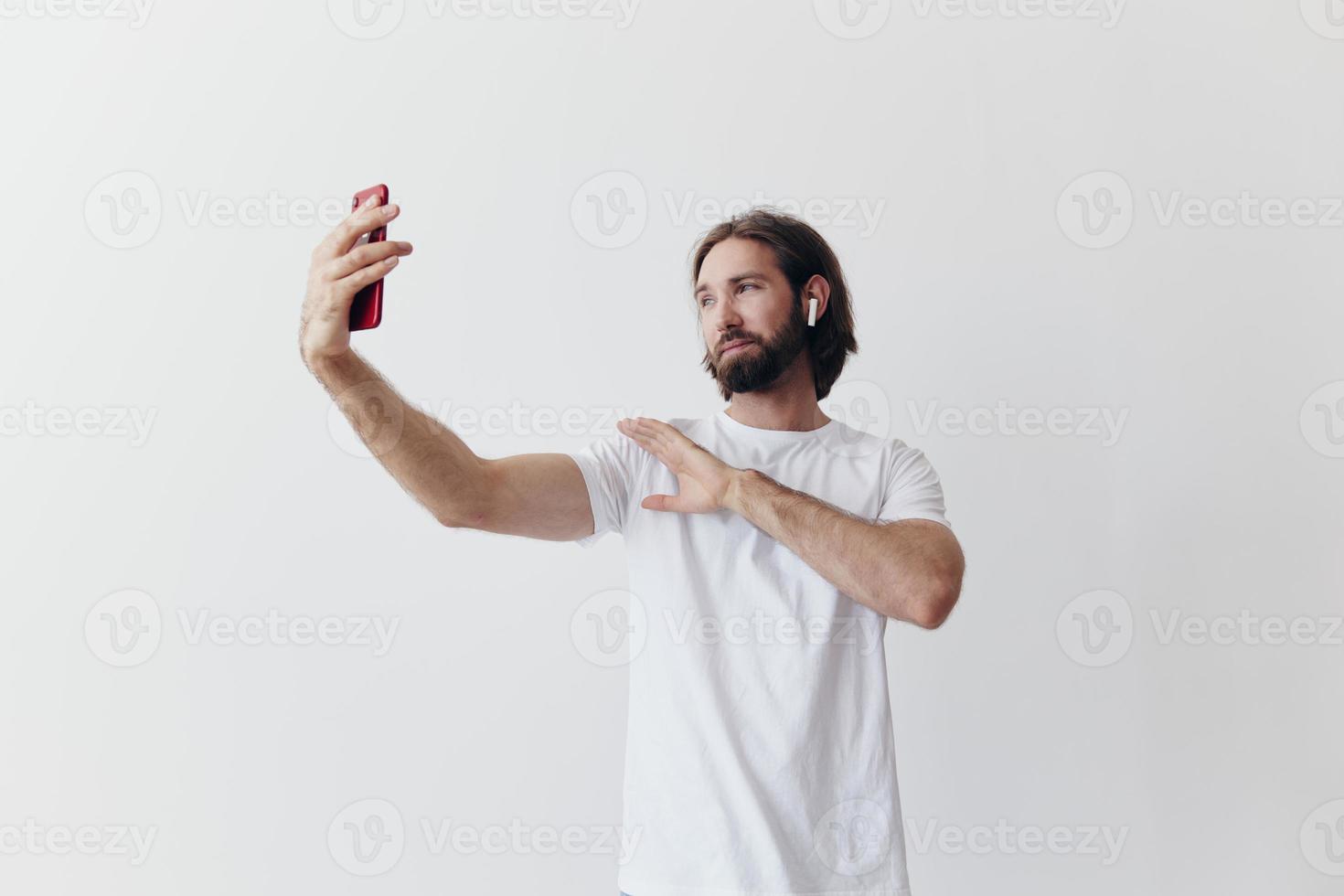 A male blogger videotaping himself on his phone and chatting with people online with a smile in a white T-shirt against a white wall photo