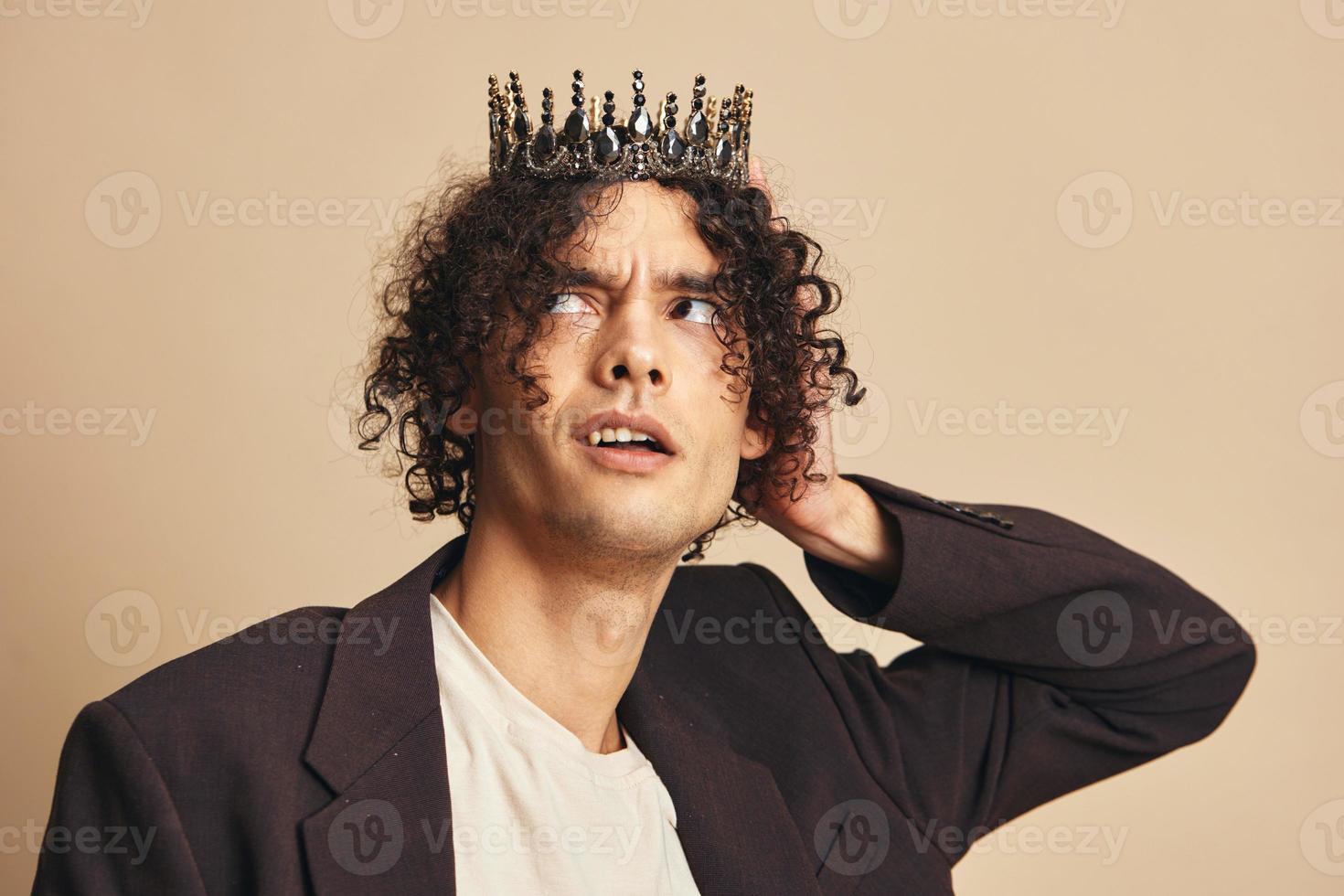Stylish unhappy handsome funny narcissistic tanned curly man with crown posing isolated on over beige pastel background. Fashion New Collection offer. Retro style concept. Free place for ad photo