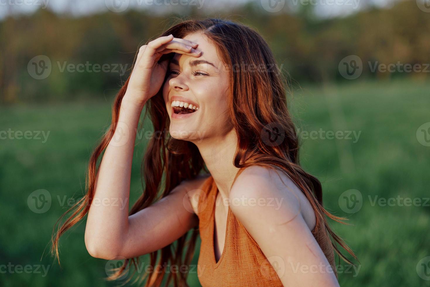 un joven mujer riendo y sonriente alegremente en naturaleza en el parque con el puesta de sol Encendiendo esclarecedor su largo rojo pelo foto