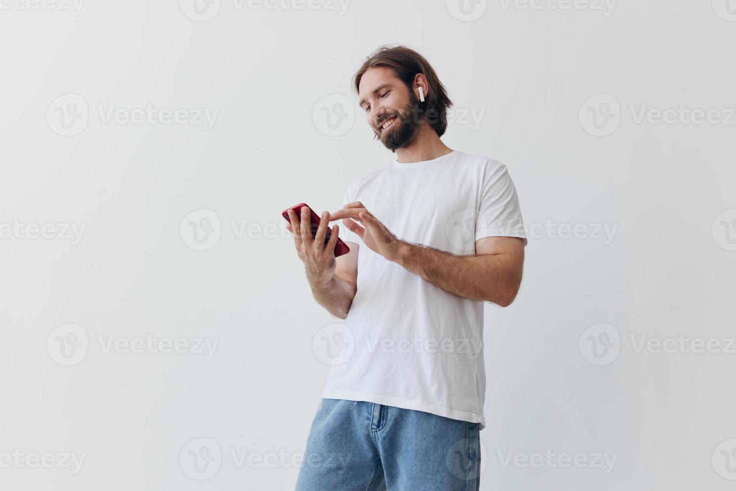 A man with a beard and long hair in a white T-shirt and blue jeans looks at his phone flipping through an online social media feed with headphones in his ears against a white background photo