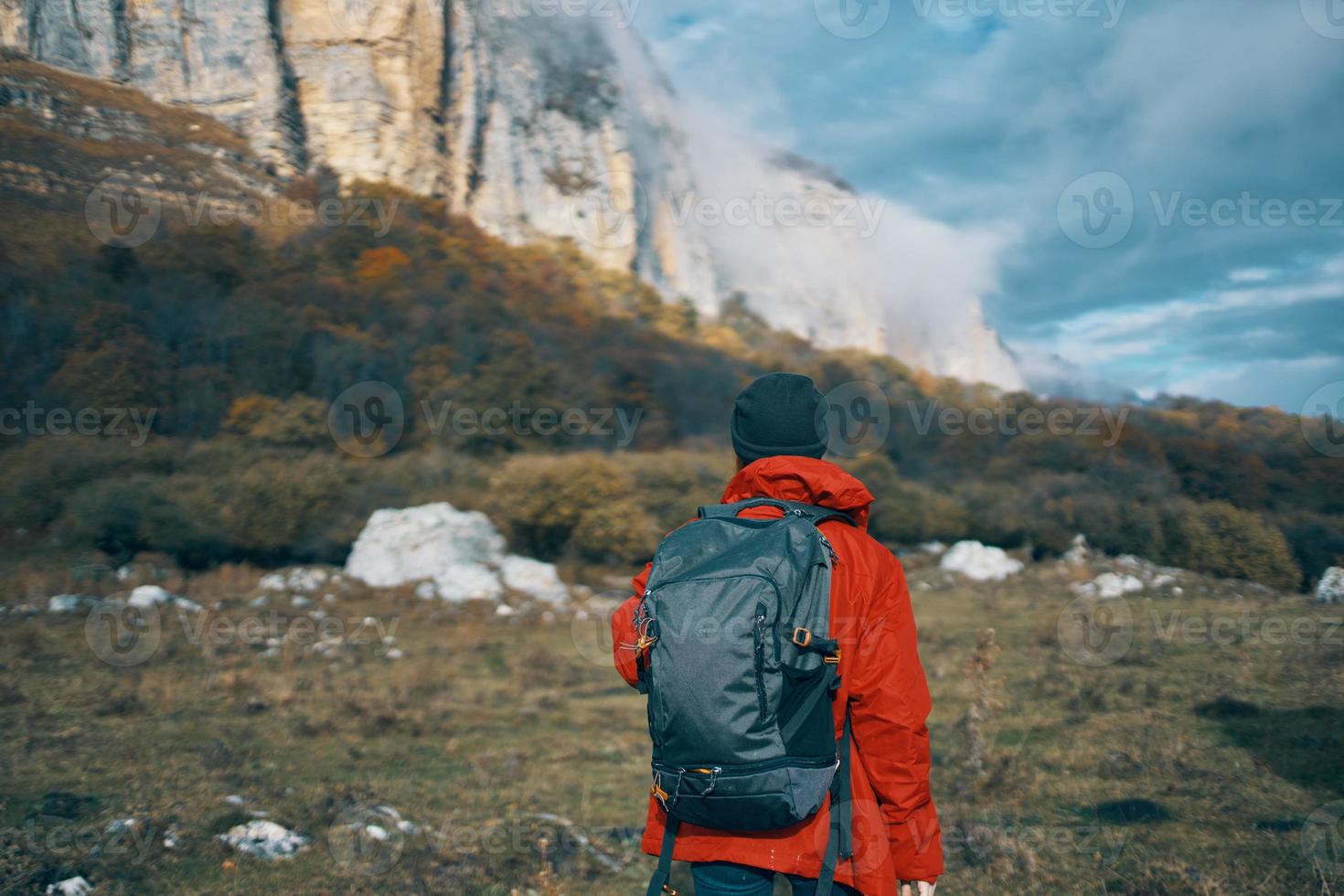 a woman in a red jacket with a backpack and in a warm hat walks on the grass in the fall in the mountains photo