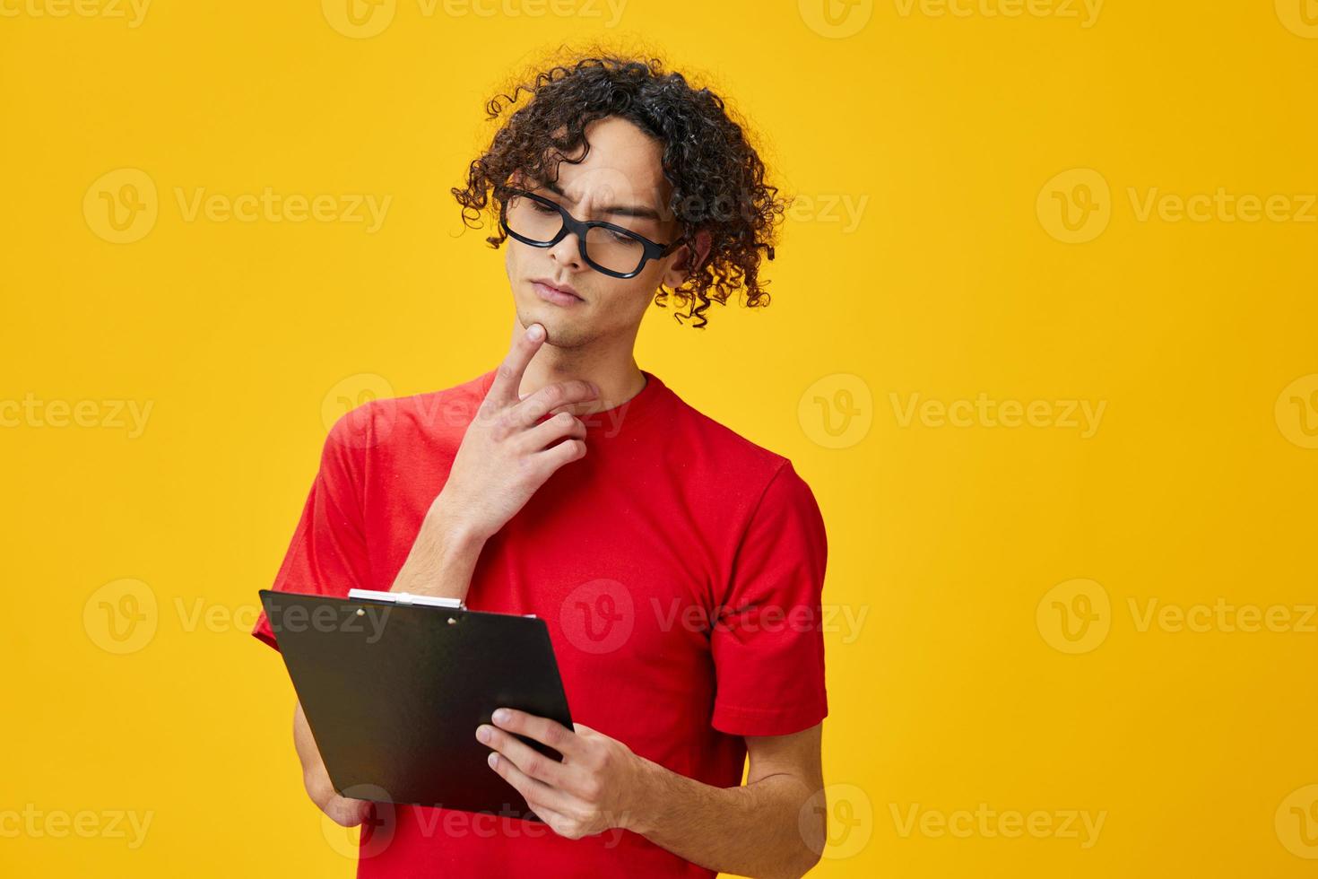 pensativo miope joven estudiante hombre en rojo camiseta gracioso gafas sostiene tableta carpeta con estudiar notas posando aislado en terminado amarillo estudio antecedentes. gratis sitio para anuncio. educación Universidad concepto foto